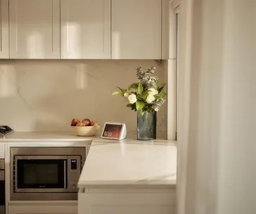 Modern kitchen with a microwave, fruit bowl, smart display showing 16:02, and a vase of white flowers on the counter.