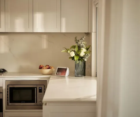 Modern kitchen with a microwave, fruit bowl, smart display showing 16:02, and a vase of white flowers on the counter.