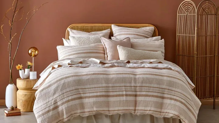 Cozy bedroom with a striped bedspread, multiple pillows, wooden side table, vase with flowers, and a rattan room divider.
