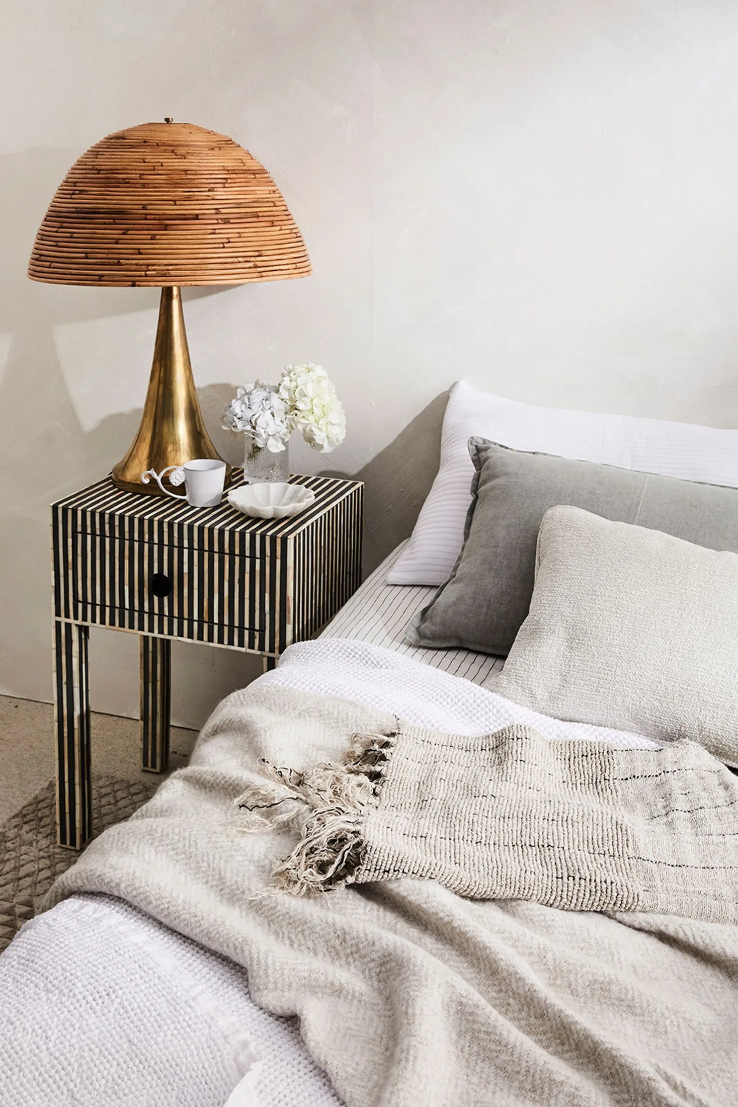 Bed with grey bed sheets, multiple blankets and three pillows beside a striped bedside table with a gold lamp