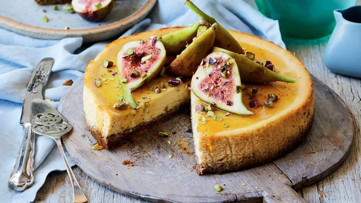 Cheesecake topped with fresh figs and chopped pistachios on a wooden board, slice removed, ornate silver serving utensils beside.