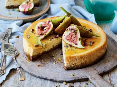 Cheesecake topped with fresh figs and chopped pistachios on a wooden board, slice removed, ornate silver serving utensils beside.