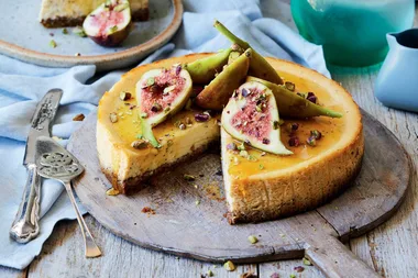 Cheesecake topped with fresh figs and chopped pistachios on a wooden board, slice removed, ornate silver serving utensils beside.