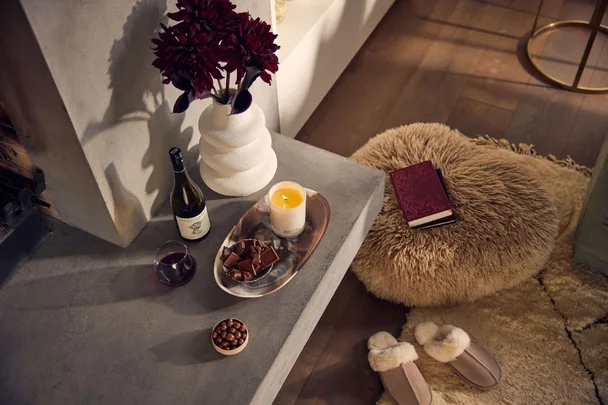 Cozy scene with wine, chocolates, a candle, red flowers, a book, fuzzy pouf, and slippers on a wood floor.