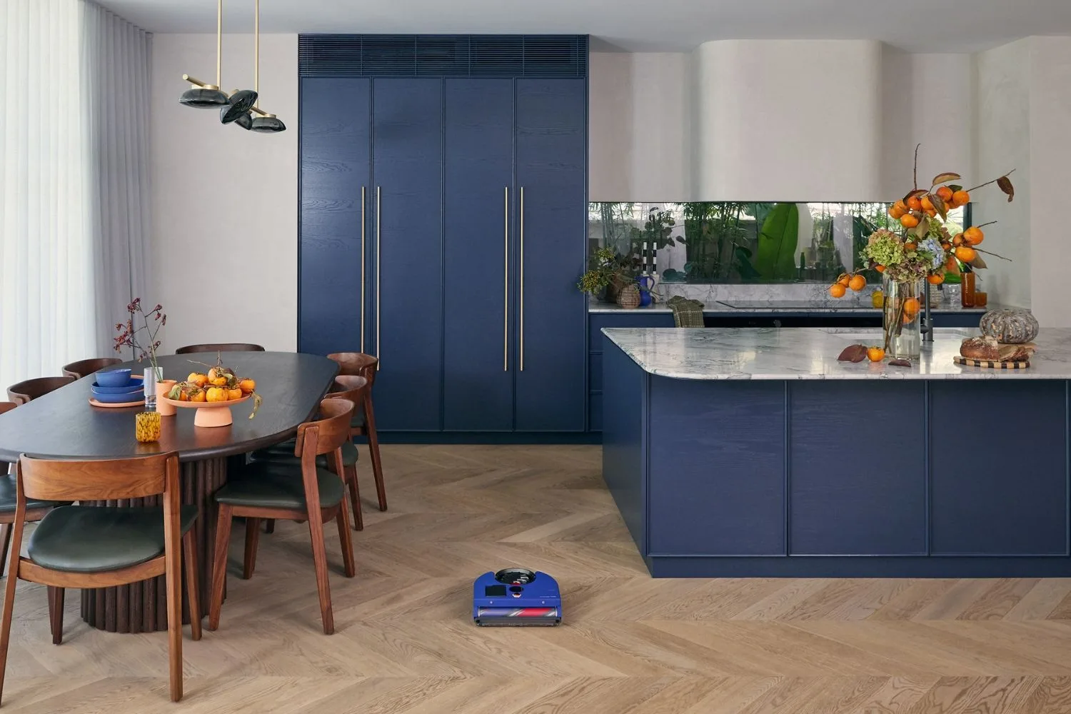 Modern kitchen with blue cabinets, wooden dining table, chairs, and a robot vacuum on the floor.