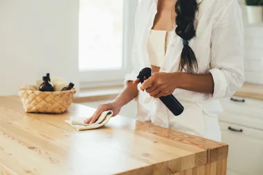 Woman using black bottle of natural homemade surface spray cleaner on a timber kitchen bench