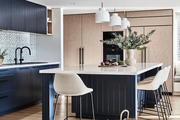 Modern kitchen with blue cabinets, patterned tile backsplash, white island, and beige bar stools under pendant lights.