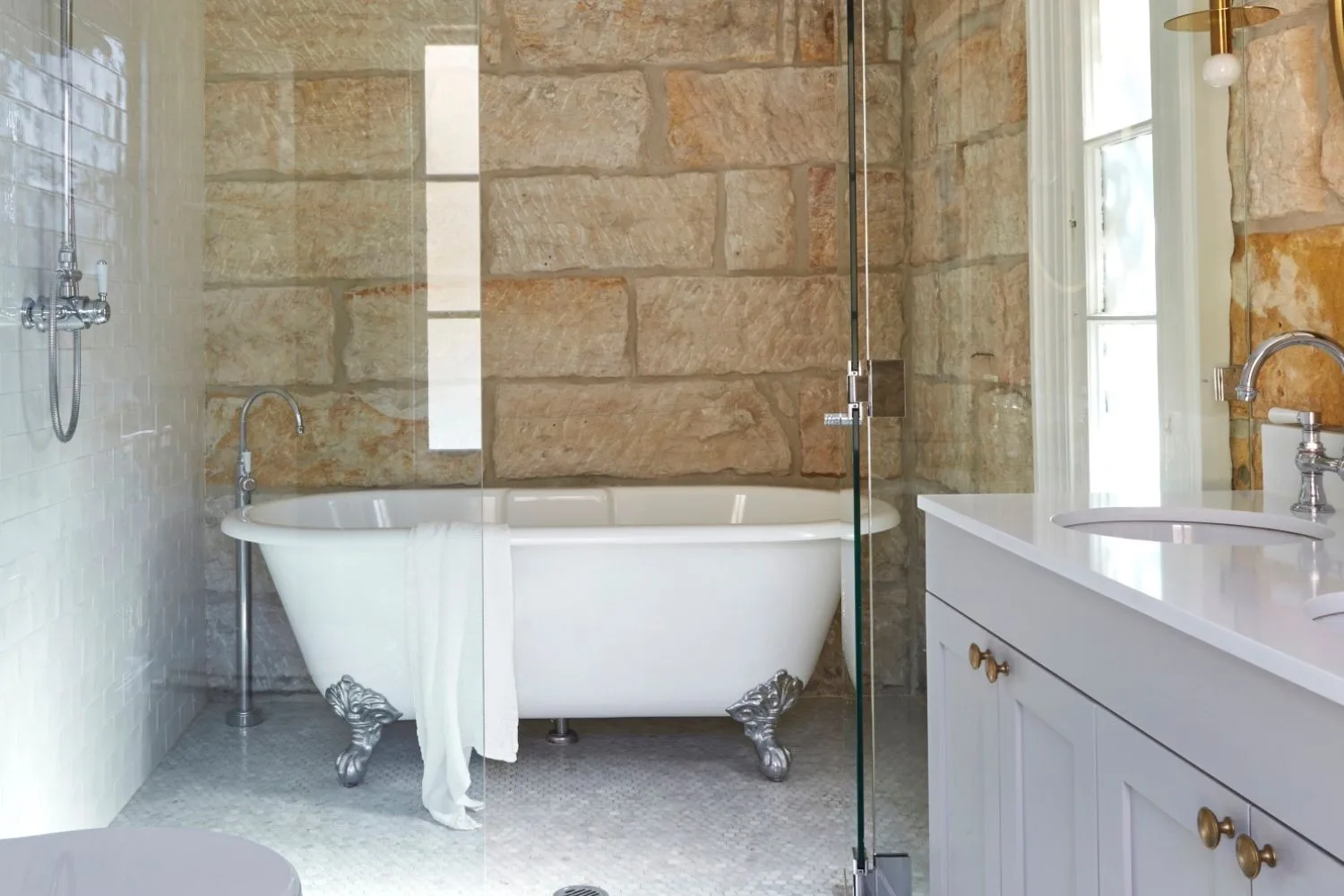 Luxurious bathroom with a clawfoot bathtub, stone wall, white vanity, glass shower, and large window.