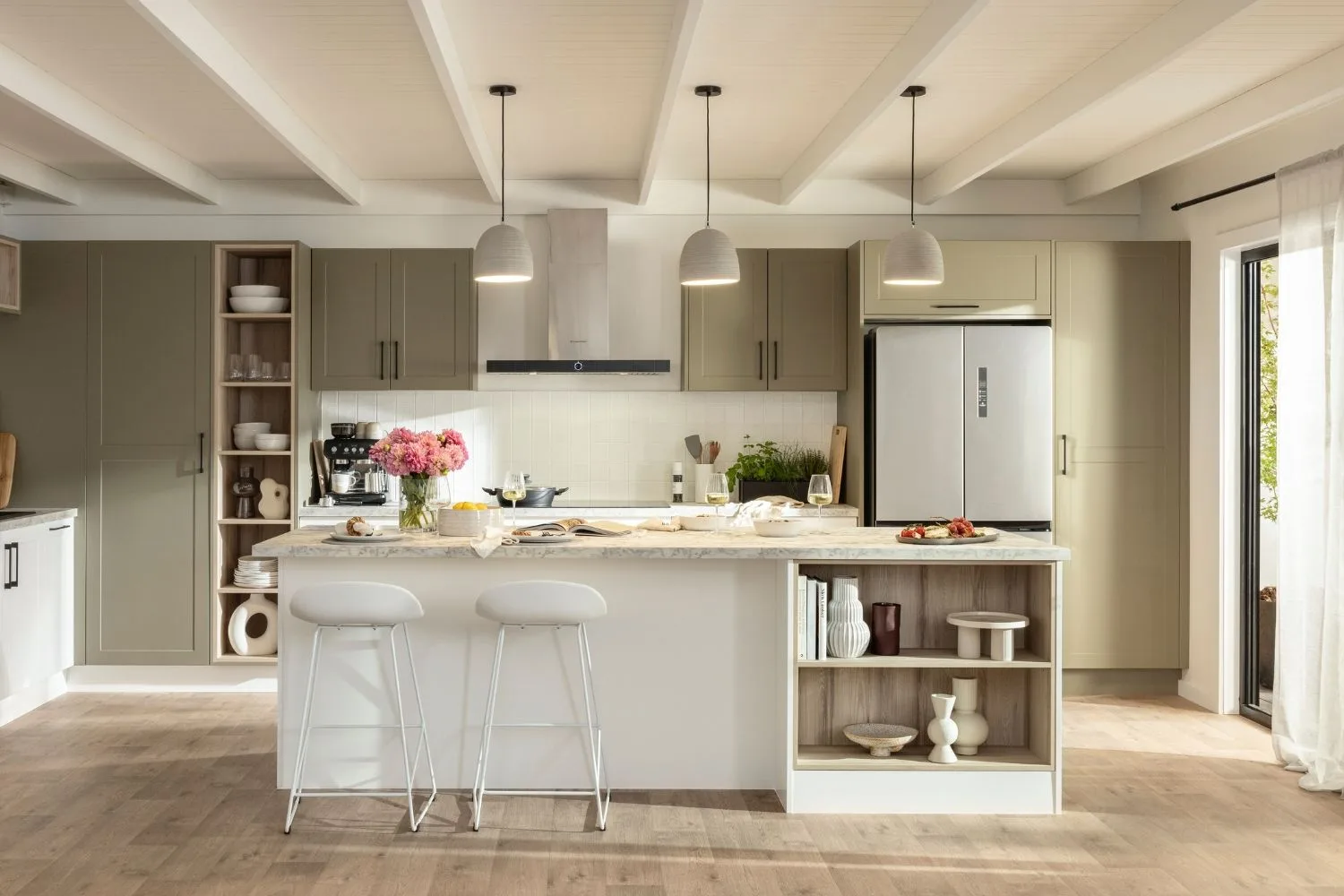 Modern kitchen with island, stools, pendant lights, and stainless steel fridge. Decor includes flowers and wine glasses.