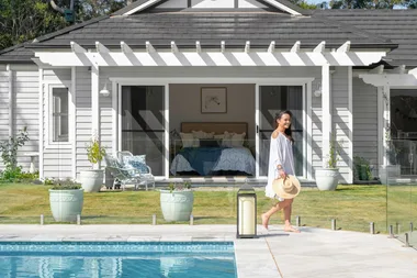 Woman walks barefoot by a pool, holding a hat, with a modern house and open sliding doors in the background.