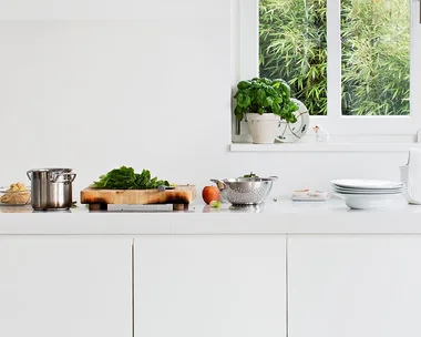 Modern white kitchen with countertop; pots, cutting board with greens, plates, and a sink by a window.