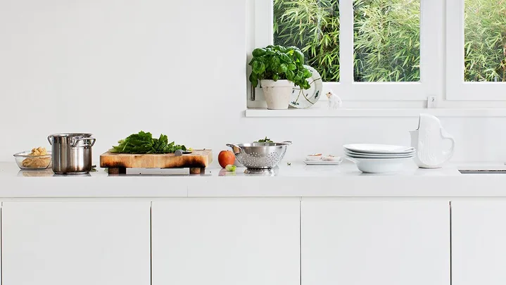 Modern white kitchen with countertop; pots, cutting board with greens, plates, and a sink by a window.