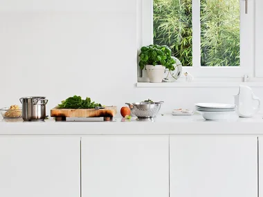 Modern white kitchen with countertop; pots, cutting board with greens, plates, and a sink by a window.