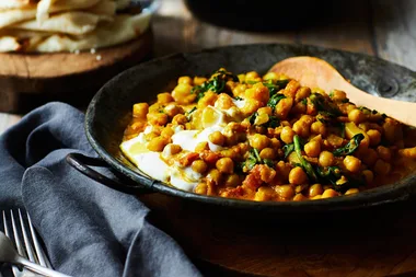 Chickpea curry with greens in a black bowl, garnished with yogurt, served beside a gray napkin and stack of flatbreads.
