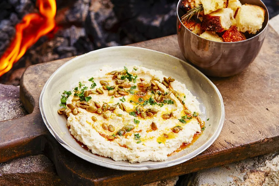 Plate of hummus with herbs and seeds on a wooden board, next to a bowl of bread cubes by a fire.