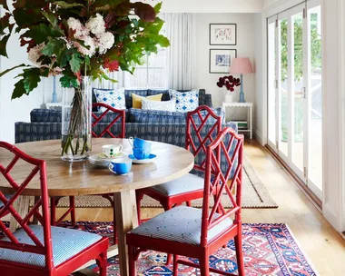Dining room with red chairs, round wooden table, floral centerpiece, blue couch, and large windows.