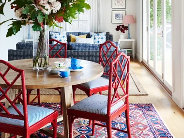 Dining room with red chairs, round wooden table, floral centerpiece, blue couch, and large windows.
