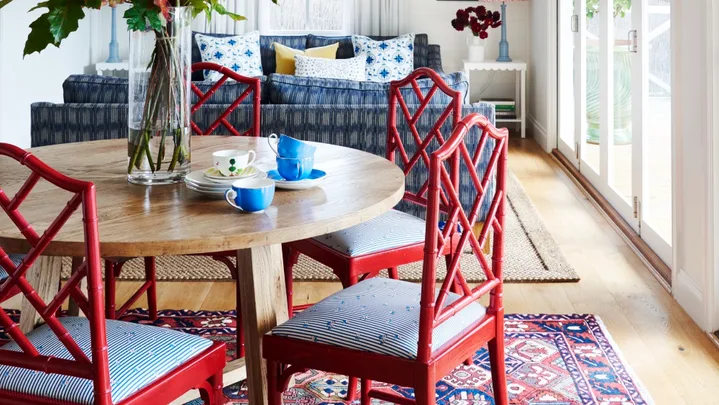 Dining room with red chairs, round wooden table, floral centerpiece, blue couch, and large windows.