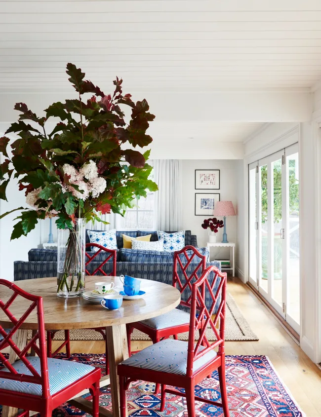 Dining room with red chairs, round wooden table, floral centerpiece, blue couch, and large windows.