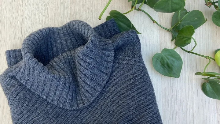 Folded gray turtleneck sweater on a light wooden surface next to green vines.