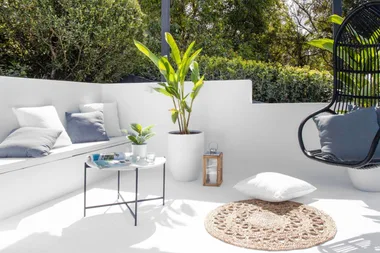 Outdoor patio with white furniture, a black swing chair, green plants, and a small table on a sunny day.
