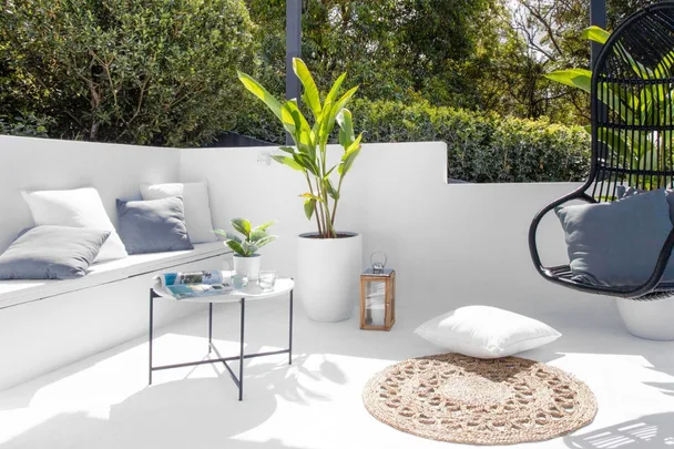 Outdoor patio with white furniture, a black swing chair, green plants, and a small table on a sunny day.