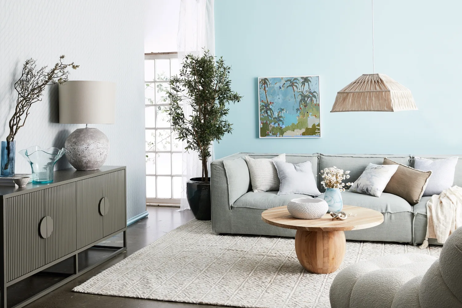 Living room with light blue walls, gray sofa, round wood table, modern decor, plants, and large window.