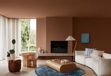 A modern living room with brown walls, white sofa, abstract chairs, a low table, and large window with view of greenery.