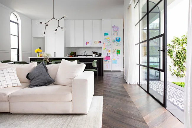Modern, open-plan living room and kitchen with a beige sofa, dynamic chandelier, and children's artwork on cabinets.