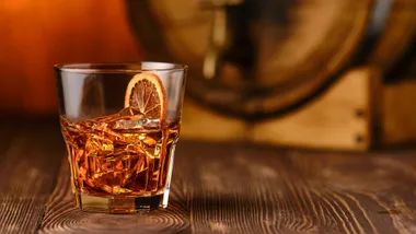 Glass of whiskey with ice and a slice of dried orange on a wooden table, barrel blurred in background.