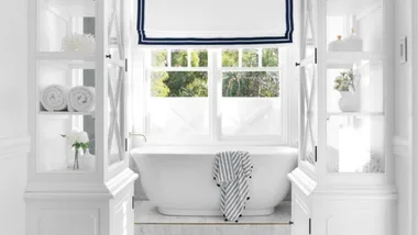 Bright white bathroom with a freestanding tub, framed by cabinets, and a window with a view of greenery outside.