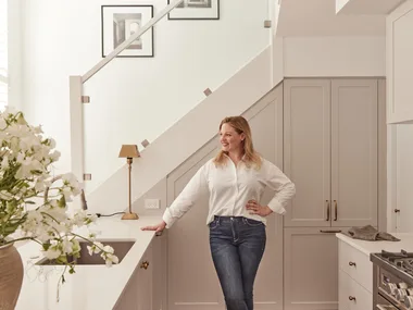Woman in a white shirt leans against a kitchen counter with flowers in the foreground and a staircase behind her.