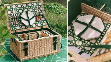 Elegant picnic basket with organized utensils, plates, and cups on a green patterned blanket outdoors.