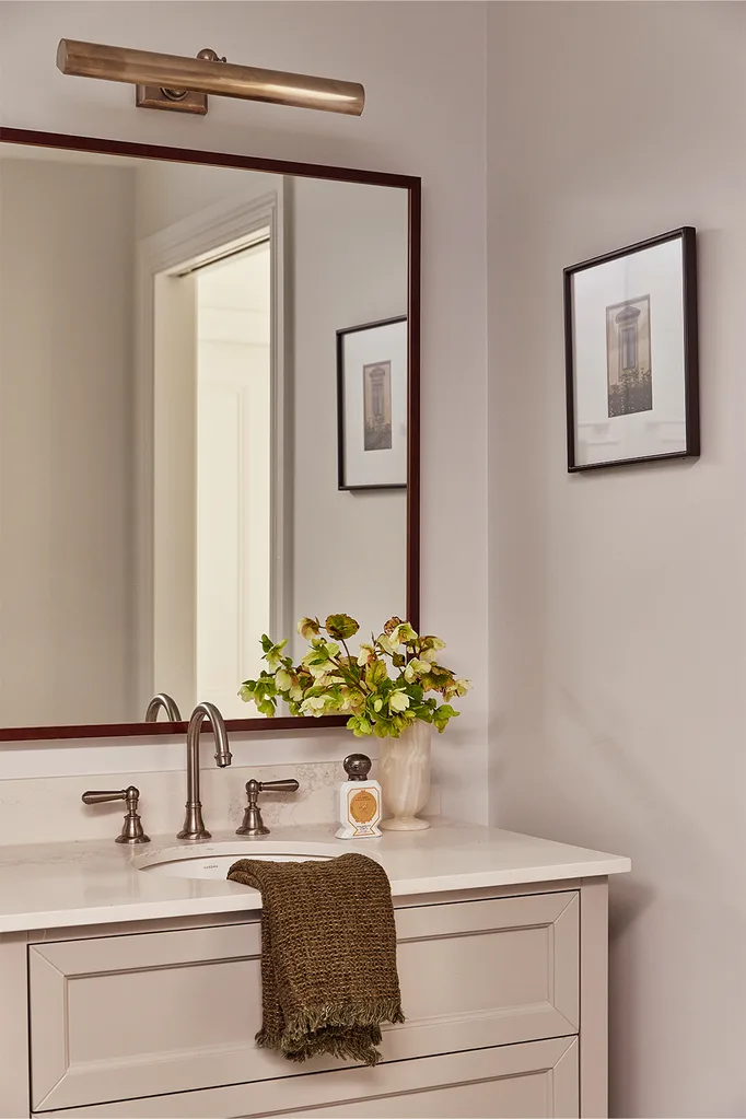 Bathroom vanity with white countertop, large mirror, brass lighting, vase with green flowers, and a brown towel.