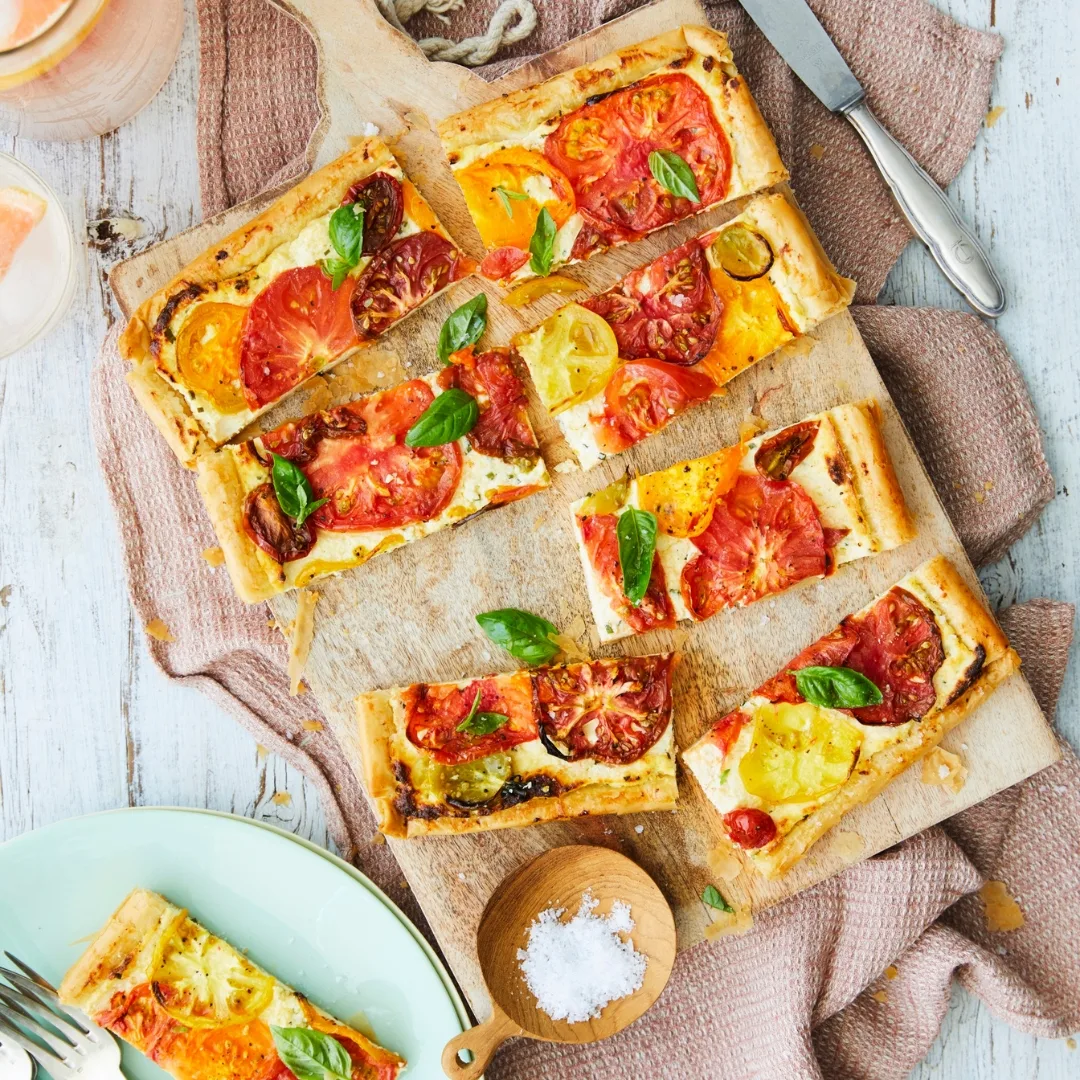 Flatbread topped with sliced tomatoes and basil on a wooden board, accompanied by a small bowl of salt and a knife.