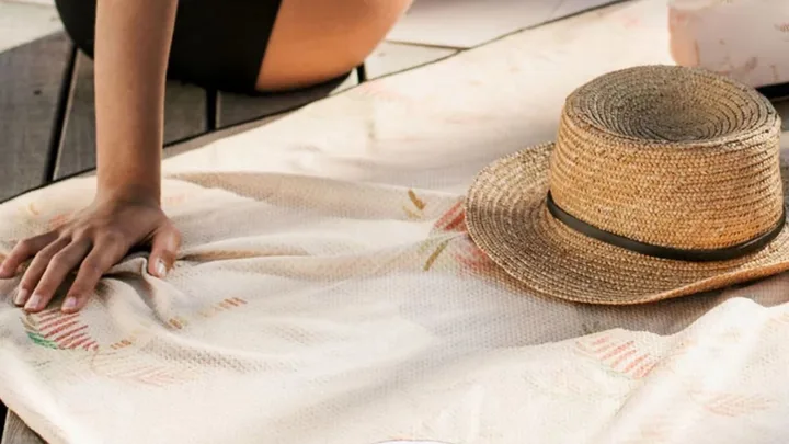 Hand on a beach towel next to a straw hat, partially seen person in a black swimsuit on a wooden deck.