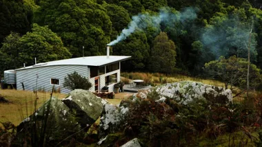 A small metal house with smoke from the chimney, surrounded by lush green forest and rocks in the foreground.