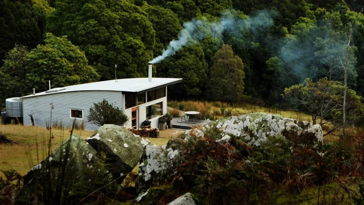 A small metal house with smoke from the chimney, surrounded by lush green forest and rocks in the foreground.