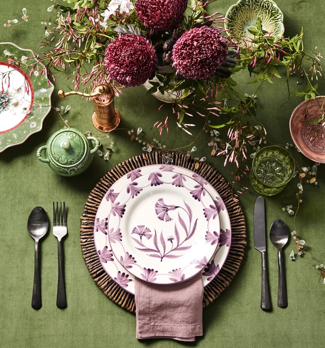 A bright green tablecloth with a vase of purple flowers and white plates with a purple floral pattern