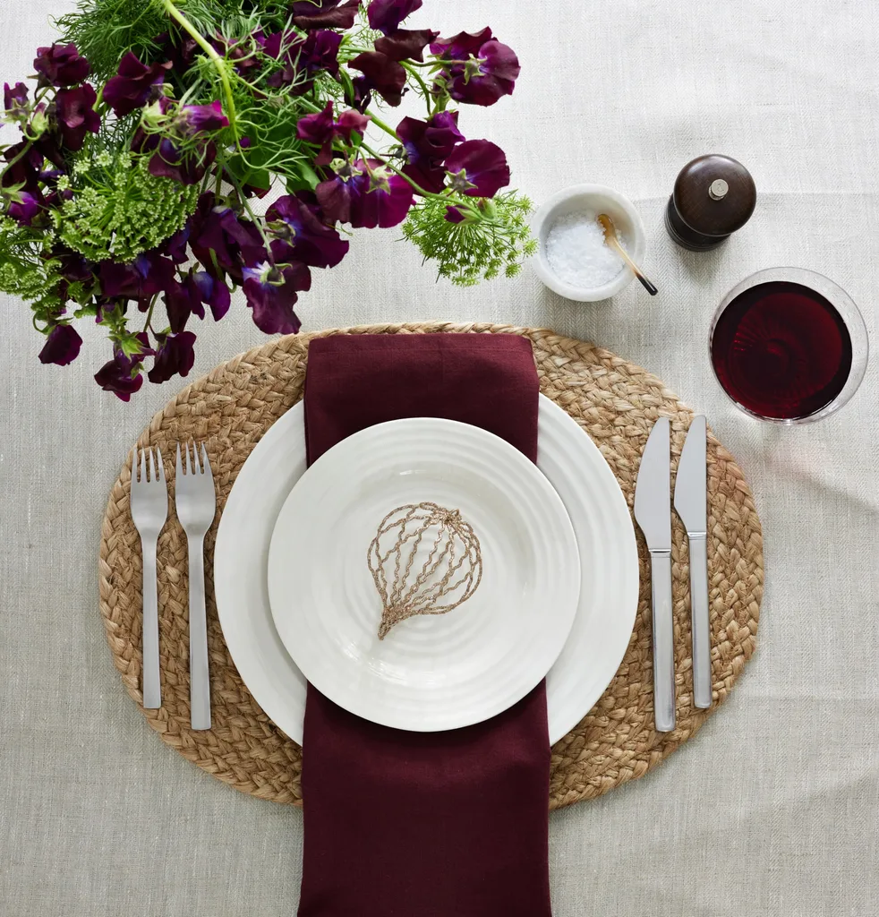 Christmas table decorations with a white tablecloth, jute placemat and a napkin in a mulled wine colour that matches the deep purple colour of the flowers arranged in a vase.
