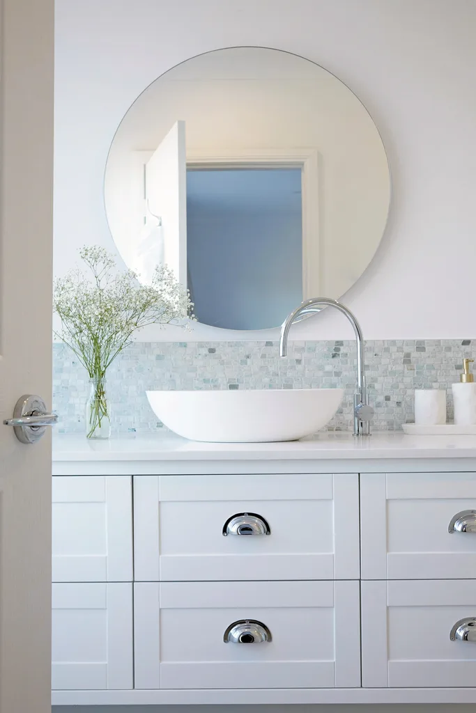 Coastal style bathroom with shaker cabinets, round mirror, mosaic tile splashback and cup handle drawer pulls.