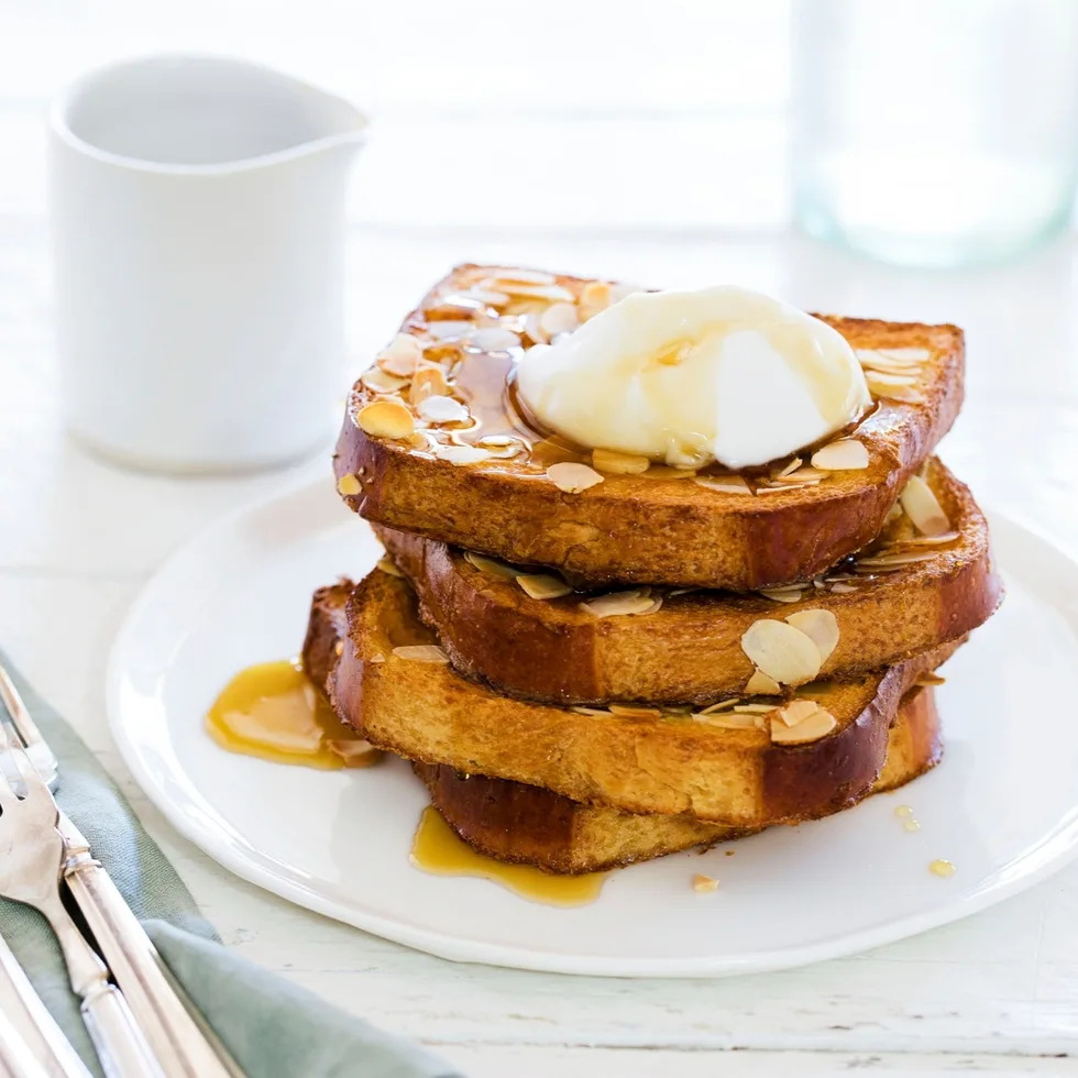 Three slices of French toast with almonds, syrup, and a dollop of cream on a white plate, with a jug in the background.
