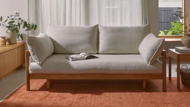 Light gray sofa with wooden frame, open book on seat, and plant on side table, in bright room with white curtains.