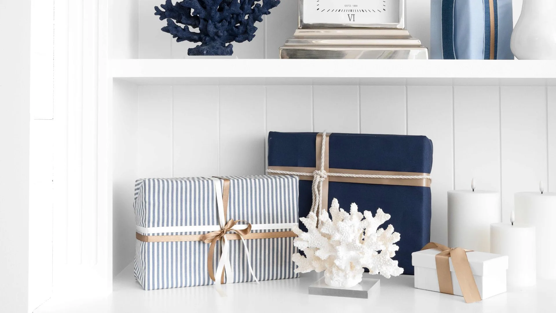 Nautical-themed gifts with blue and striped wrapping, coral decor, and candles on a white shelf.