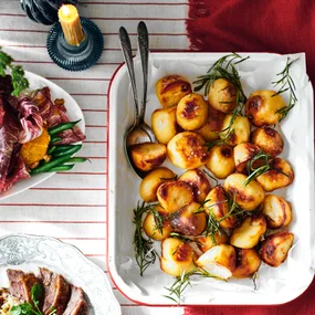 Roasted potatoes with rosemary on a tray beside a salad plate with greens and orange slices on a striped tablecloth.