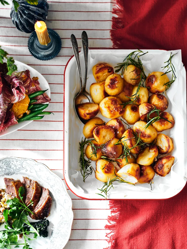 Roast potatoes in an enamel dish on a table with a red runner and red-striped tablecloth.