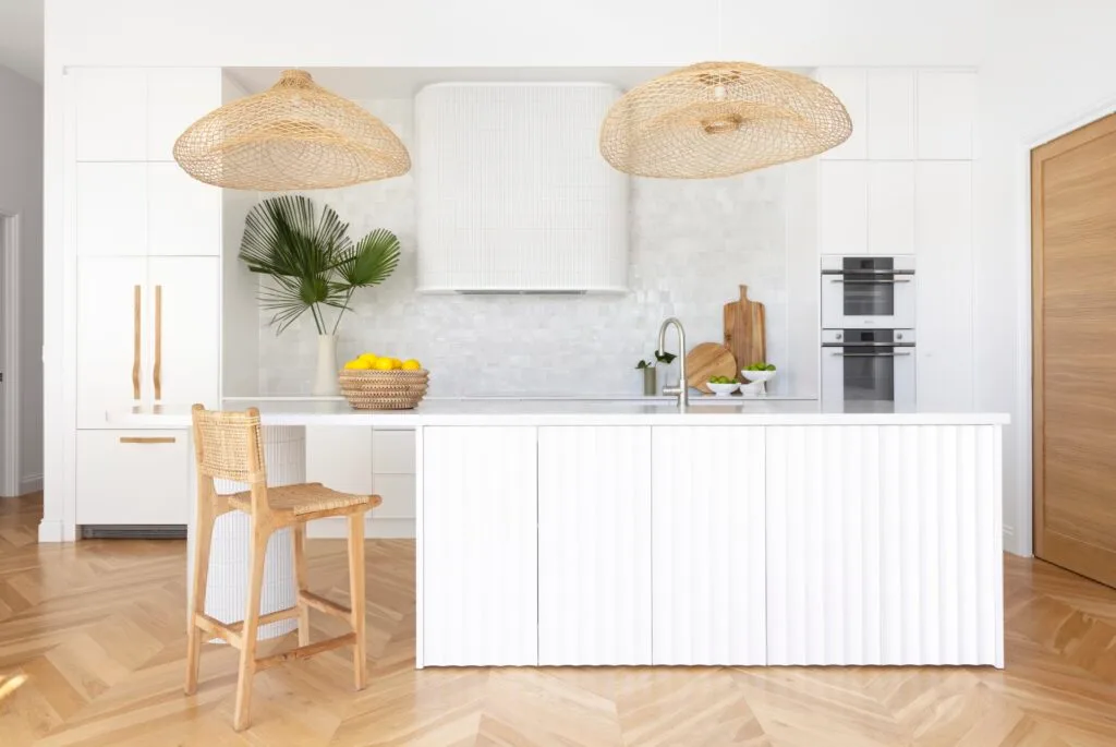 A white resort-style kitchen with woven hanging pendants and a fluted kitchen island.