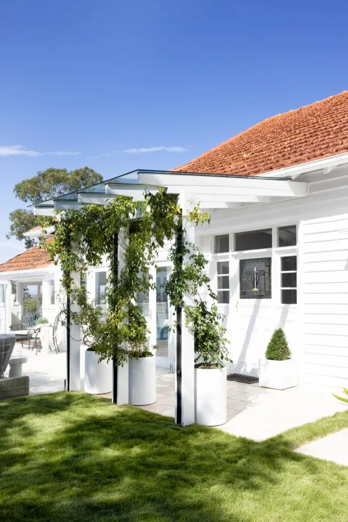 A white timber cladded bungalow with an arbour covered with creeping plants.