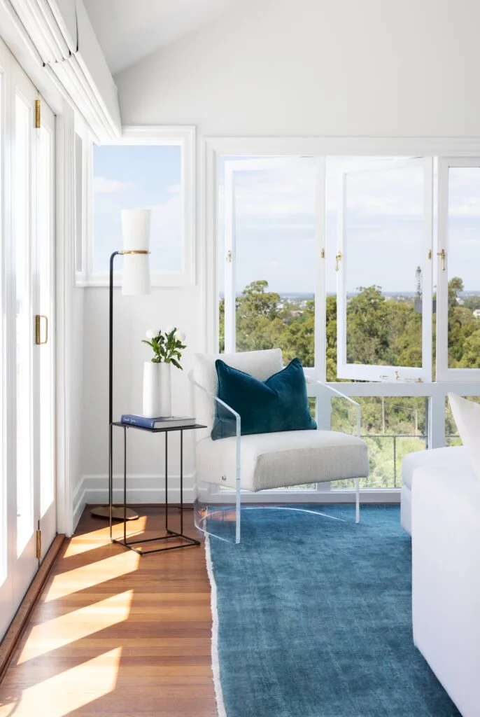 A white boucle armchair with a blue cushion beside open windows.