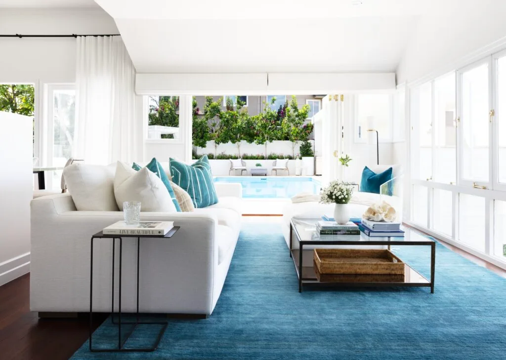 A classic white living area with a white sofa and blue rug looking out onto the pool.